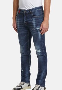 Hombre vistiendo jeans oscuros desgastados con manchas de pintura y zapatillas blancas, de pie contra un fondo liso.