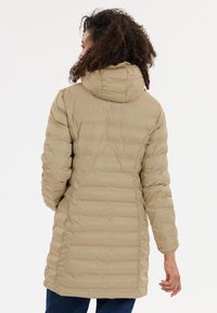 Kvinde med krøllet hår if�ørt en beige quiltet hættejakke, stående med ryggen til mod en helt hvid baggrund.