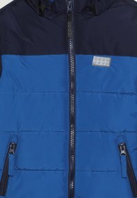 Ocieplana kurtka o granatowo-niebieskim wzorze z blokami kolorów, z centralnym zamkiem błyskawicznym, dwoma zamkami błyskawicznymi po bokach oraz naszywką z logo na piersi.