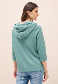 Türkise Kapuzenjacke mit Dreiviertelärmeln, aus geripptem Stoff gefertigt. Verfügt über eine lockere Passform und ein schlichtes Design. Getragen mit hellblauen Jeans.
