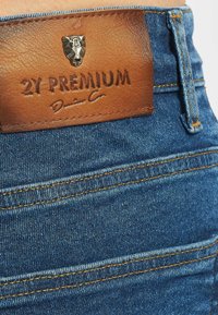 Denim-farkut, joissa on ruskea nahkalappu, johon on painettu "2Y PREMIUM". Vaaleansininen kangas, jossa on kultaiset tikkaukset ja lähikuva vyötärönauhasta.