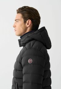 Črna napihljiva jakna s kapuco, ki ima prešito zasnovo, gladko teksturo in okroglo oznako logotipa na levem ramenu.