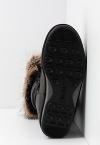 Botas de invierno negras con ribete de piel de pelo, mostrando una suela gruesa con un patrón de tracción circular y la marca "MOON BOOT", en ángulo contra paredes blancas.