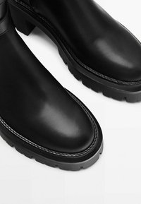 Bottes en cuir noir avec des bouts ronds et des semelles épaisses et rainurées, présentées en gros plan sur un fond blanc.
