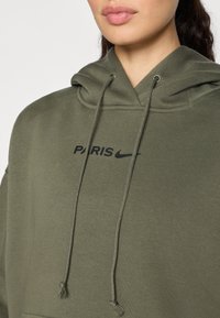 Sudadera de color verde oliva hecha de una tela suave que presenta un logo negro de "PARIS" y el swoosh de Nike, con cordones ajustables y un acabado texturizado.