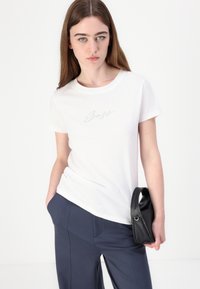 White cotton t-shirt with a crew neck and short sleeves, featuring silver script embroidery on the chest. Paired with dark blue pants and a black bag.