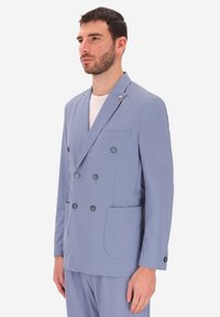 Uomo che indossa un blazer doppiopetto blu chiaro con bottoni neri e pantaloni abbinati, camicia bianca sotto, su uno sfondo bianco semplice.