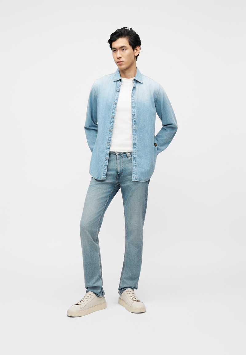 Junger Mann steht mit den Händen hinter dem Rücken, trägt ein hellblaues Jeanshemd, weißes T-Shirt, hellblaue Jeans und weiße Sneakers auf weißem Hintergrund.