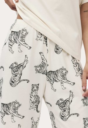 Person trägt weiße Hose mit schwarz umrandeten Tigerillustrationen in verschiedenen Posen und ein schlichtes, hellbeiges Oberteil.