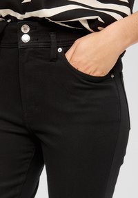 Zwarte jeans hebben een slim fit met een gladde textuur, hoge tailleband en opvallende zilveren knopen. De zakken zijn subtiel geïntegreerd.