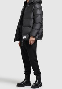 Czarna pikowana kurtka z kapturem, kieszeniami na suwak i elastycznymi mankietami, połączona z czarnymi spodniami dresowymi i masywnymi czarnymi butami.