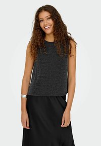Czarny top bez rękawów z fakturowanym, wzorzystym wzorem, w połączeniu z gładką czarną spódnicą. Modelka nosi srebrną bransoletkę mankietową.