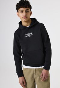 Sudadera negra con un bolsillo frontal, capucha y texto blanco que dice "PERSPECTIVA FUTURA". Lleva puesta sobre una camiseta blanca y combinada con pantalones beige.