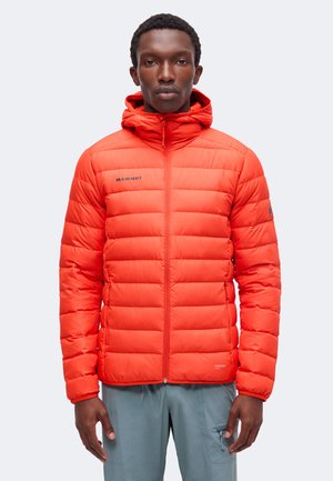 Man die een feloranje doorgestikte kapmantel draagt met ritszakken en een grijze broek, staand tegen een effen lichte achtergrond.