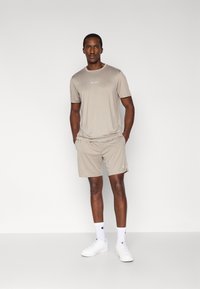 Αθλητική μπλούζα μπεζ και ταιριαστά σορτς από ελαφρύ ύφασμα. Διαθέτουν μικρό λογότυπο στο στήθος και κλασική εφαρμογή. Λευκά αθλητικά παπούτσια και κάλτσες.