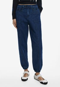 Pantalones jogger de denim azul oscuro con puños elásticos, bolsillos abotonados y un corte relajado. Llevados con zapatos slip-on blancos y negros.