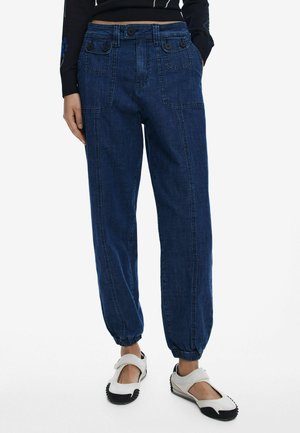 Dark blue denim jogger pants with elastic cuffs, buttoned pockets, and a relaxed fit. Worn with white and black slip-on shoes.