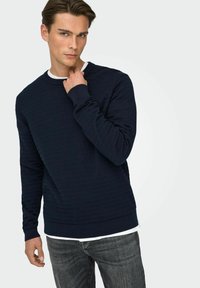 Sweatshirt bleu marine texturé avec poignets et ourlet côtelés, présentant un col ras du cou. Porté sur une chemise blanche, associé à un jean foncé.