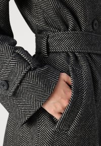 Abrigo de patrón espiga en gris y blanco con cinturón, que presenta puños abotonados y un bolsillo lateral. Tejido de lana texturizado.