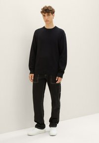 Maglione nero a maniche lunghe con tessuto strutturato, abbinato a jeans neri larghi con cuciture bianche a contrasto e sneakers bianche.
