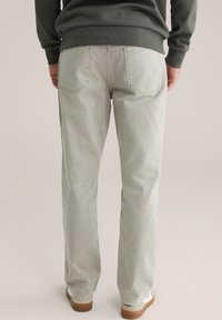 Hombre que lleva vaqueros rectos de color gris claro, sudadera verde oscuro y zapatillas blancas con suelas marrones, de pie de espaldas.
