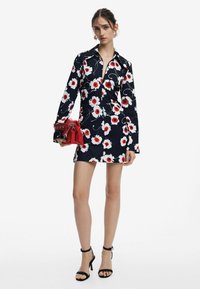 Abito camicia floreale con sfondo blu navy, caratterizzato da grandi fiori rossi e bianchi. Maniche lunghe con polsini abbottonati. Tieni una borsa rossa.