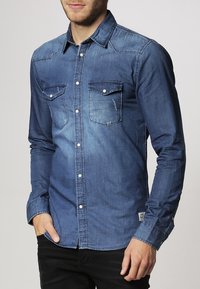 Tummansininen pitkähihaisen farkkupaita, jossa on kaksi rintataskua, nappikiinnitys ja tikattu koristelu. Pehmeä kangas, jossa haalistunut kuvio.