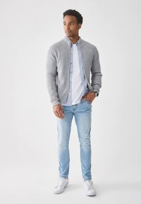 Szürke bordázott kardigán világoskék gombos ing fölött, világoskék farmerrel és fehér tornacipővel párosítva; laza, réteges ruha.