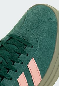 Gros plan d'une basket en daim vert avec des lacets vert foncé et deux bandes en cuir rose sur le côté avec une semelle en caoutchouc beige.
