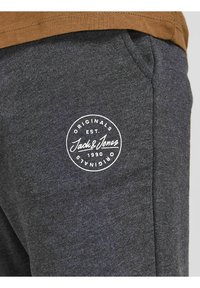 Šedé tepláky vyrobené z měkké tkaniny, s kulatou bílou nášivkou na stehně s textem "Jack & Jones" a datem založení.