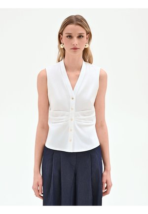 Femme aux longs cheveux blonds porte un haut blanc sans manches boutonné et un pantalon plissé bleu marine sur un fond clair uni.