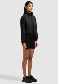 Ženska stoji oblečena v črno jopico z zadrgo, črne kolesarske kratke hlače in črne športne čevlje na enobarvnem svetlo sivem ozadju.