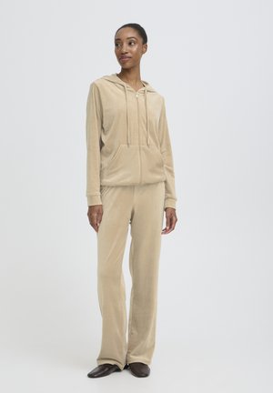 Vrouw die staat en een beige velours hoodie met rits en bijpassende wijde broek draagt, met donkerbruine platte schoenen op een effen lichte achtergrond.