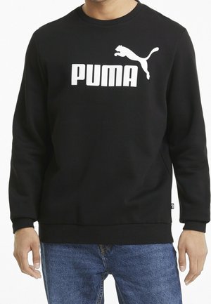 Mężczyzna nosi czarną bluzę z białym skaczącym logo pumy oraz napisem "PUMA", w połączeniu z niebieskimi dżinsami.