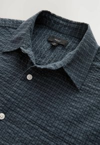 Button-up skjorte i mørkeblå med et struktureret, ternet mønster, der har en krave og hvide knapper. Brandetiket synlig inden i nakkeområdet.