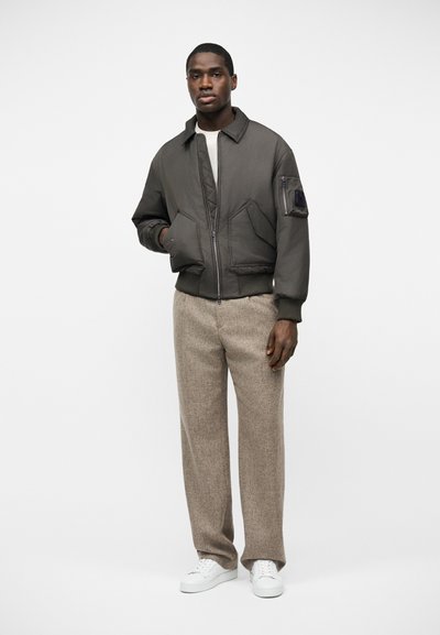 Homme portant une veste bomber zippée vert foncé, un pantalon large beige et des baskets blanches, debout devant un fond blanc uni.