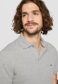 Grå polo skjorte med teksturert overflate, krage med to knapper, og liten trekantet brodering med røde og blå detaljer.