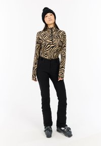 Pantaloni da sci neri aderenti abbinati a una maglia a maniche lunghe con zip, decorata con una stampa zebrada marrone e nera. Berretto lavorato a maglia nero e scarponi da sci.