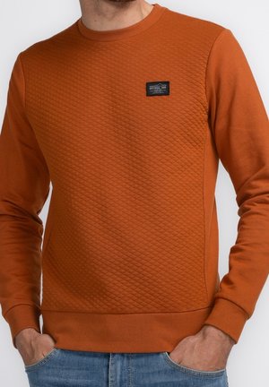 Mand iført rust-orange quiltet langærmet trøje med sort rektangulært mærke på brystet og blå jeans, hænderne i lommerne.