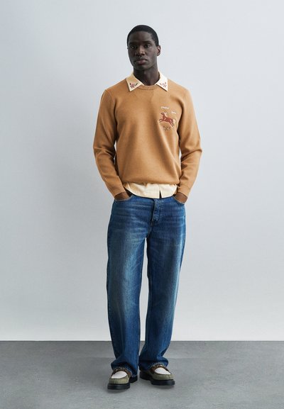 Άνδρας που στέκεται με τα χέρια στις τσέπες, φοράει πουλόβερ σε ταμπ χρώμα με κέντημα αλόγου, εκρού πουκάμισο με γιακά, μπλε τζιν και δίχρωμα μοκασίνια.