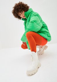 Person with curly hair squatting, wearing a bright green puffer jacket, orange leggings, and white chunky boots against a plain background.