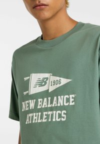 Grøn bomulds t-shirt med korte ærmer, der har et hvidt grafisk design af et flag og "NEW BALANCE ATHLETICS 1906" i fed skrift.