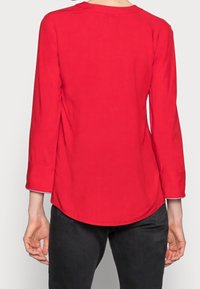 Blusa rossa a maniche lunghe con orlo arrotondato, caratterizzata da un colletto semplice e piping bianco sui polsini, realizzata in tessuto leggero.