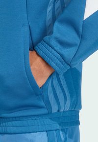 Modrá športová bunda má hladkú textúru, rebrované manžety a skrytú vrecko. Dizajn obsahuje kontrastné modré bočné pruhy.