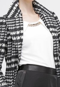 Černobílý kabát s vzorem psího zubu a širokým límcem, nošený přes bílý top s detaily zlatého řetězu, doplněný černým opaskem.