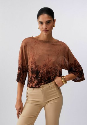 Donna con capelli pettinati all'indietro che indossa orecchini a cerchio dorati, una maglia floreale marrone trasparente con maniche a gomito e pantaloni beige a vita alta.