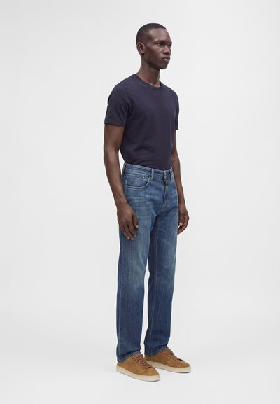 Homme debout portant un t-shirt bleu marine foncé, un jean bleu et des baskets beige sur un fond gris clair uni.