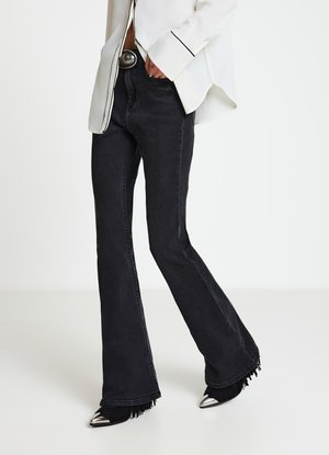 Jeans neri a zampa d'elefante con orlo sfrangiato, realizzati in denim, abbinati a tacchi con punta a punta. Il modello indossa una camicia bianca con piping nero.