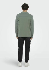 Sudadera verde con cuello, mangas largas y detalle de costura en la parte trasera, combinada con pantalones cargo negros y zapatillas blancas.