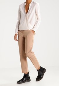 Bílá blůza s dlouhými rukávy a výstřihem do V, spárovaná s elegantními béžovými kalhotami a černými lakovanými kotníkovými botami s texturovaným povrchem.